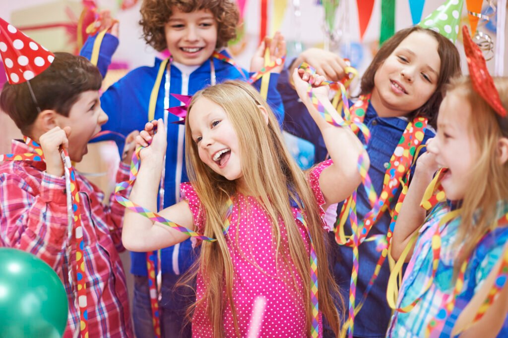 Fröhliche Kinder in bunten Faschingskostümen tanzen und feiern gemeinsam, umgeben von Luftballons und Konfetti.