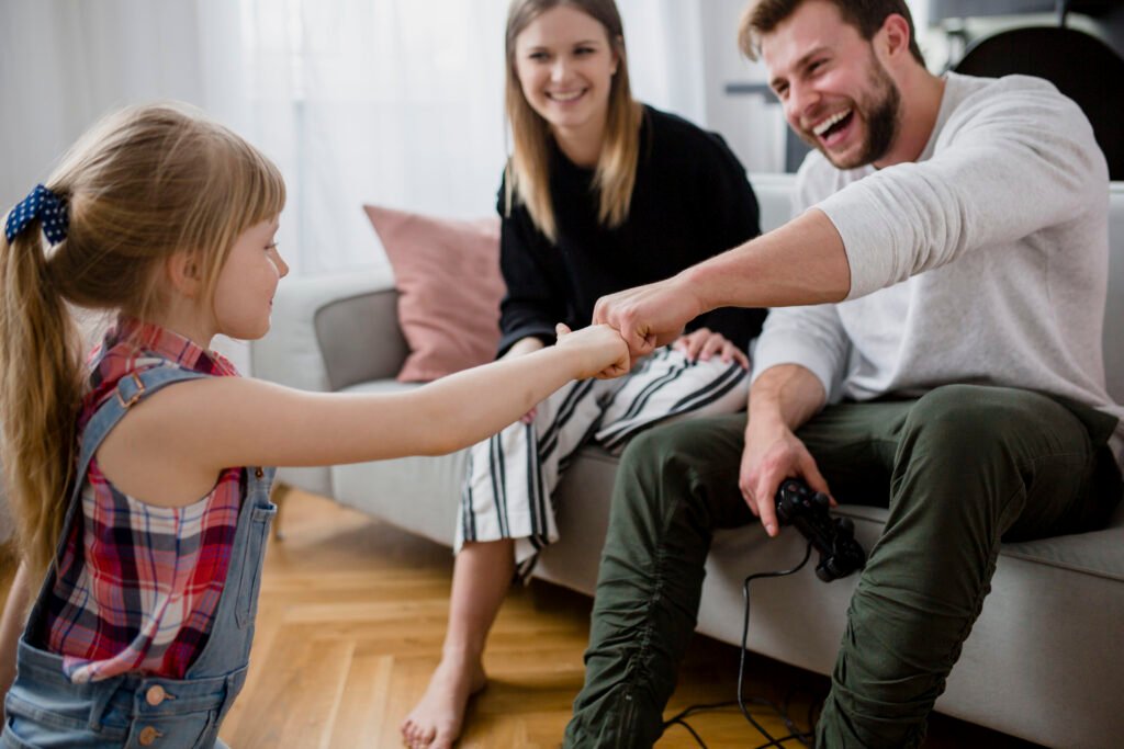 Ein Kind und sein Papa geben sich fröhlich einen Hand-Check, während die Mama daneben sitzt und lächelnd zuschaut. Die Szene zeigt eine liebevolle Familienbindung und positive Erziehungsmomente.