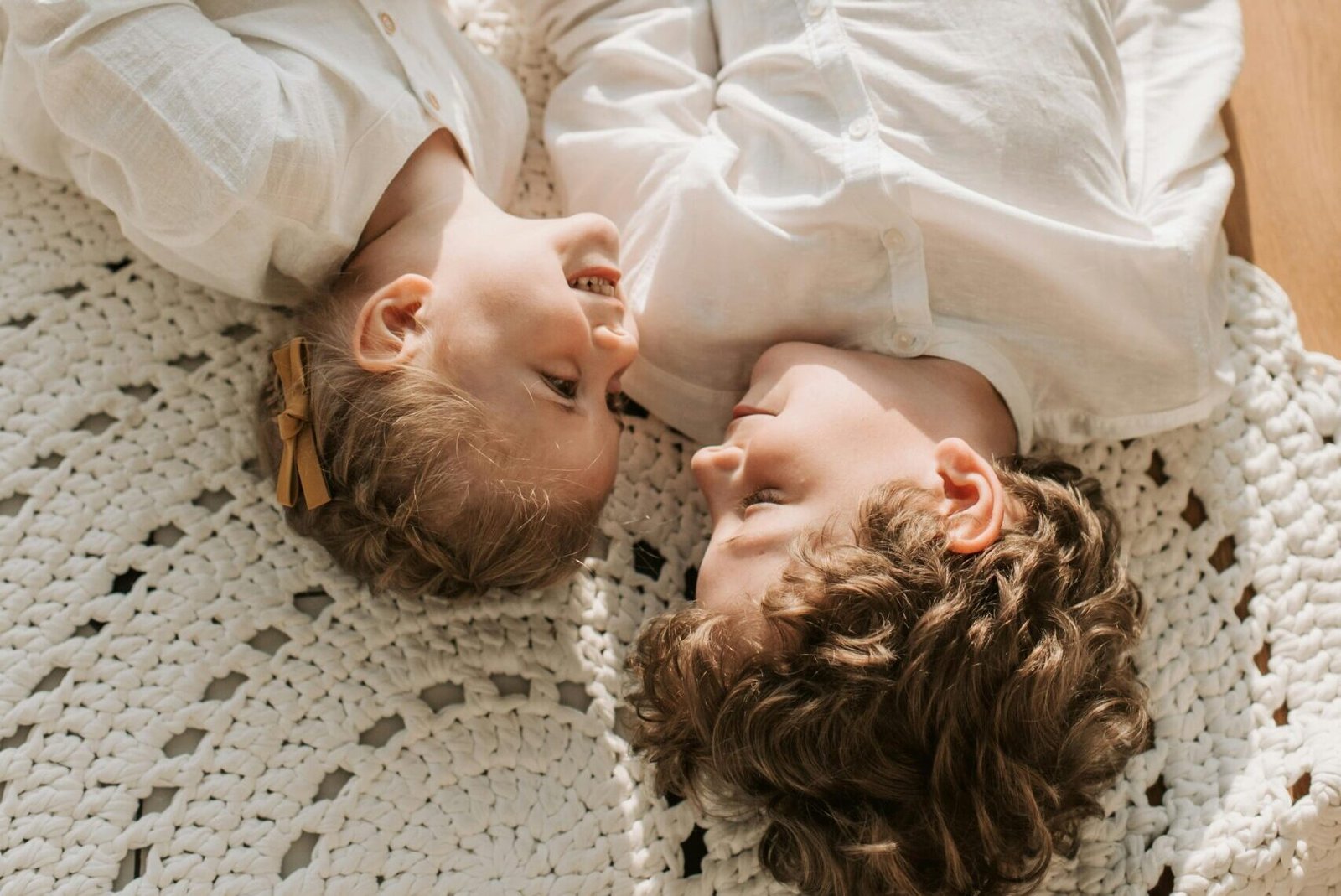 Zwei Kinder sitzen zusammen auf dem Boden, eines etwas älter, eines jünger. Die ältere Schwester schaut liebevoll auf den kleinen Bruder, während beide gemeinsam ein Buch anschauen.